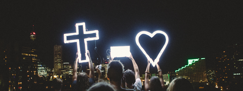 Young people holding a glowing cross and heart at night in front of a city skyline